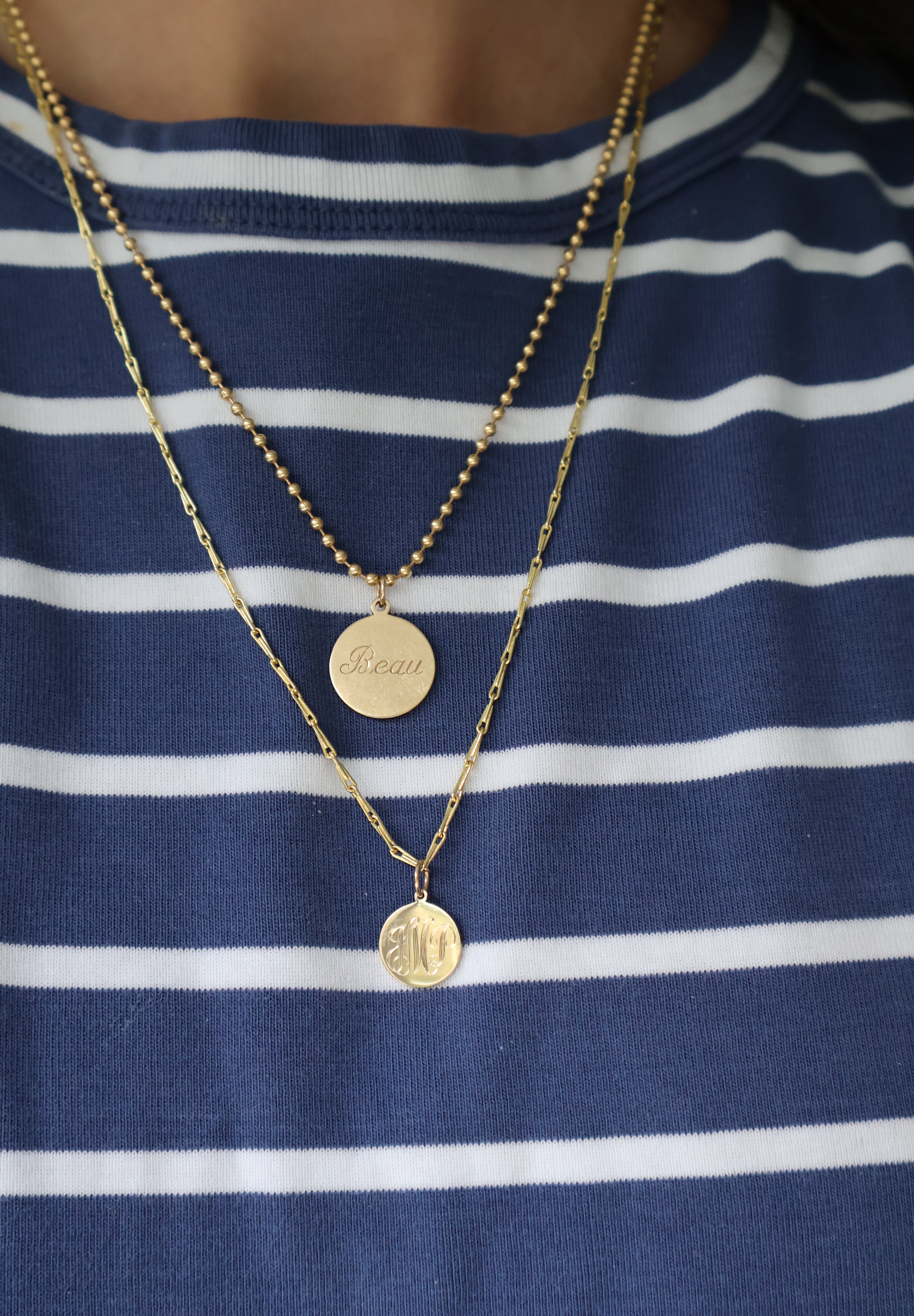 Two layered gold necklaces worn by a person wearing a blue and white striped shirt. The top necklace has a small round disc pendant, and the bottom necklace has a larger circular pendant with an engraved symbol.