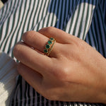 A gold ring with three green emerald-cut stones worn on a person's hand, resting on a green and white striped fabric background.