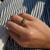 A gold ring with three green emerald-cut stones worn on a person's hand, resting on a green and white striped fabric background.