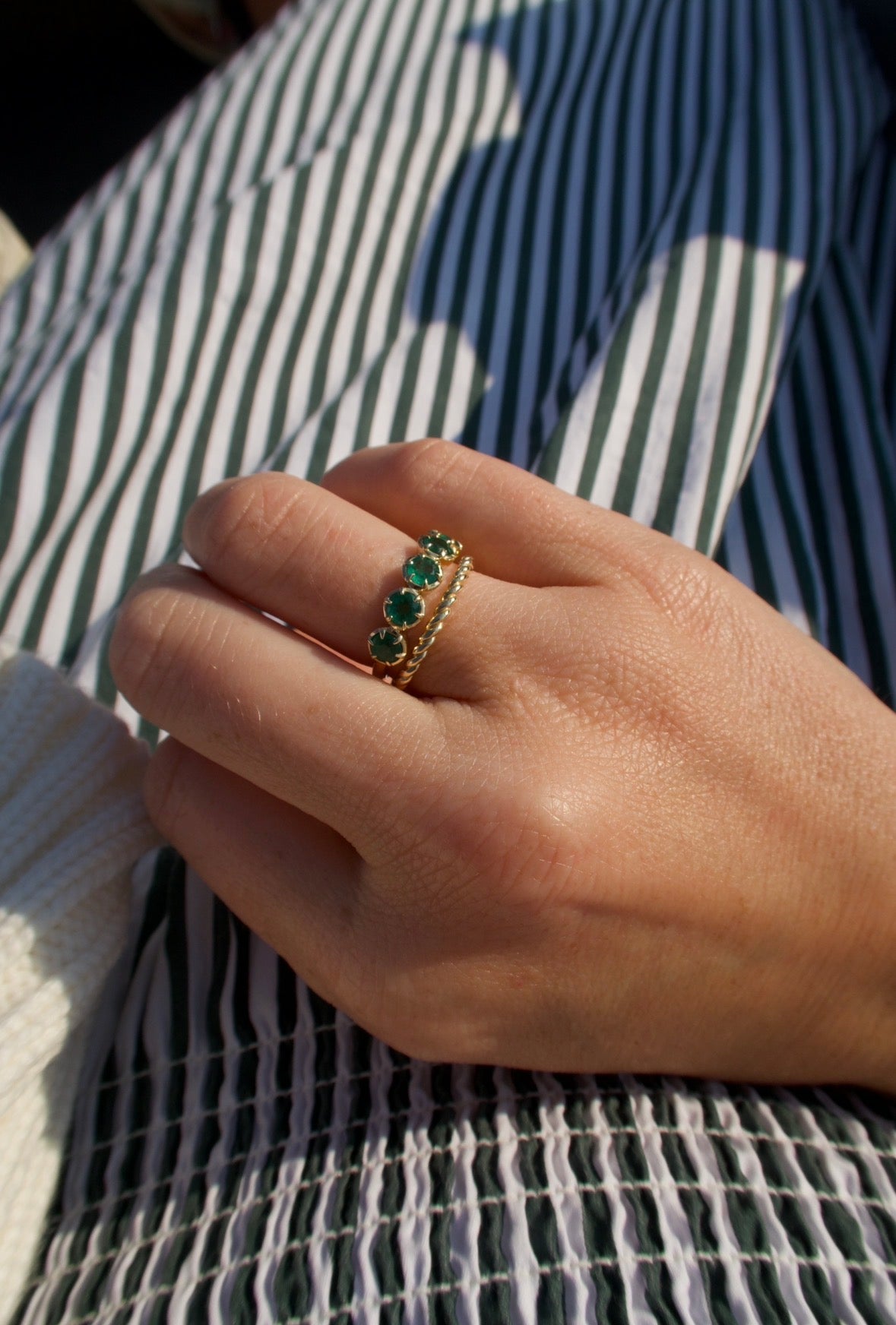 A gold ring with three green emerald-cut stones worn on a person's hand, resting on a green and white striped fabric background.