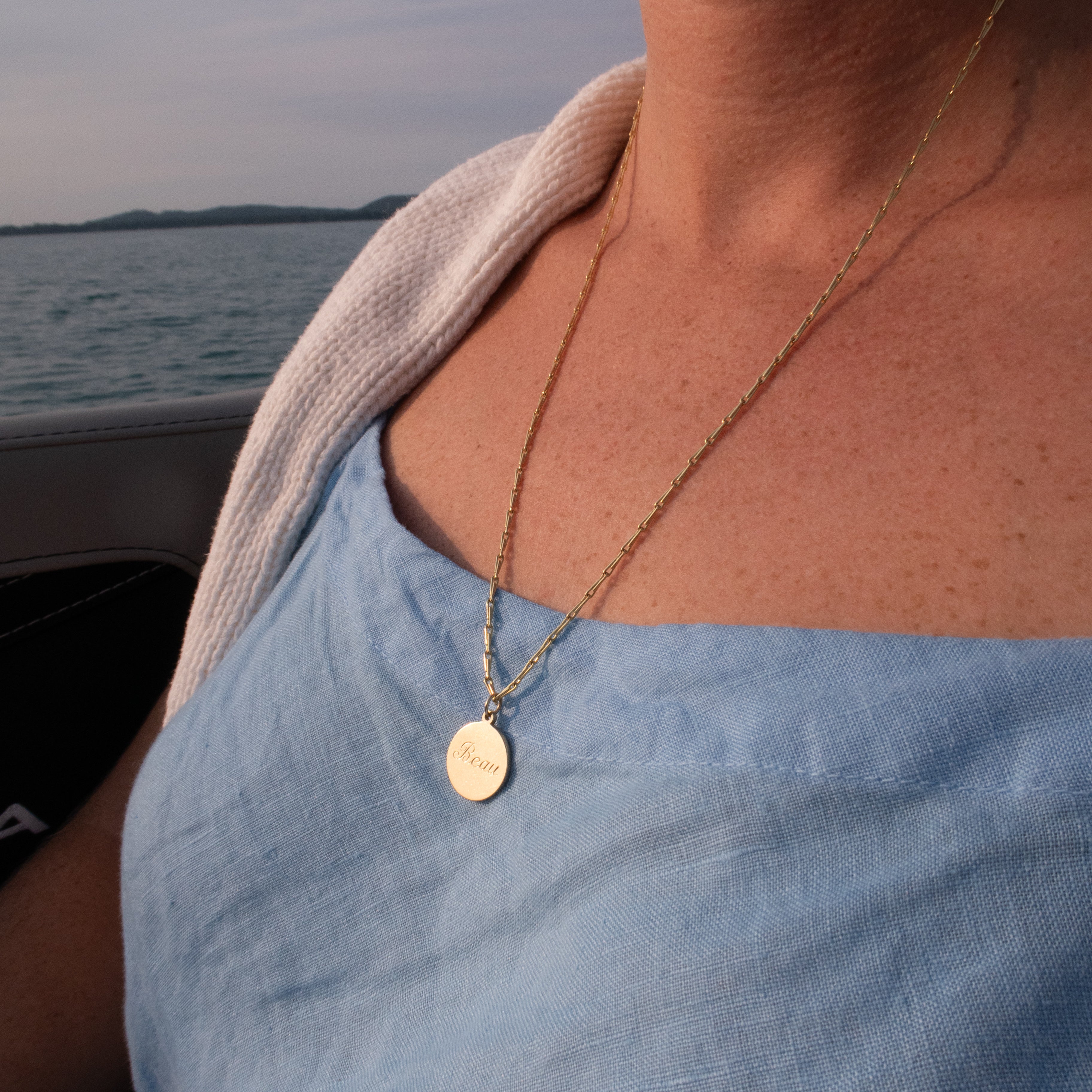 A close-up lifestyle shot of a woman wearing a delicate gold chain necklace with a round, flat gold pendant, set against a background of water and sky.