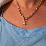 A close-up lifestyle shot of a person wearing a gold chain necklace with a small clear gemstone pendant, styled with a light blue top and a beige knit sweater.