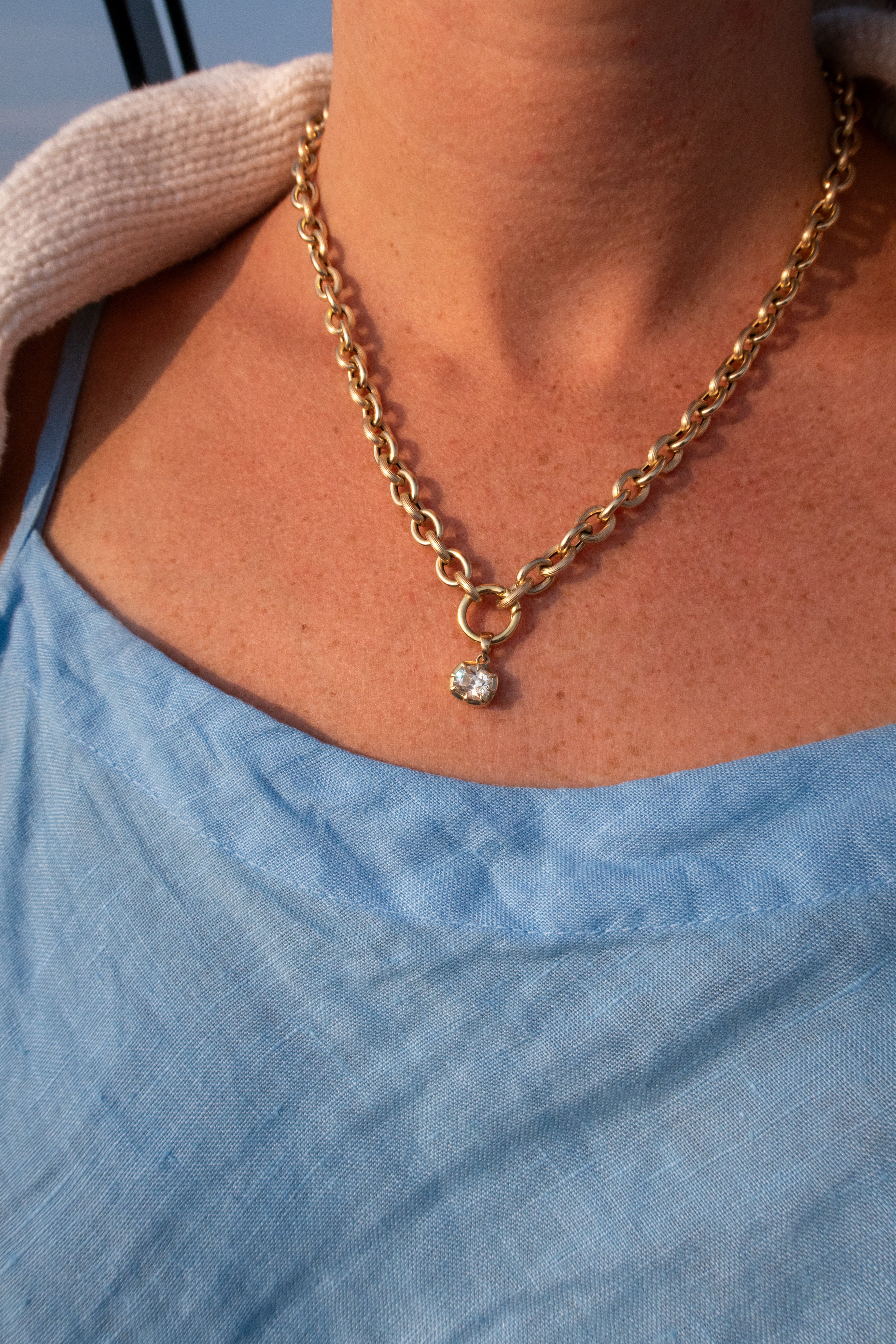 A close-up lifestyle shot of a person wearing a gold chain necklace with a small clear gemstone pendant, styled with a light blue top and a beige knit sweater.