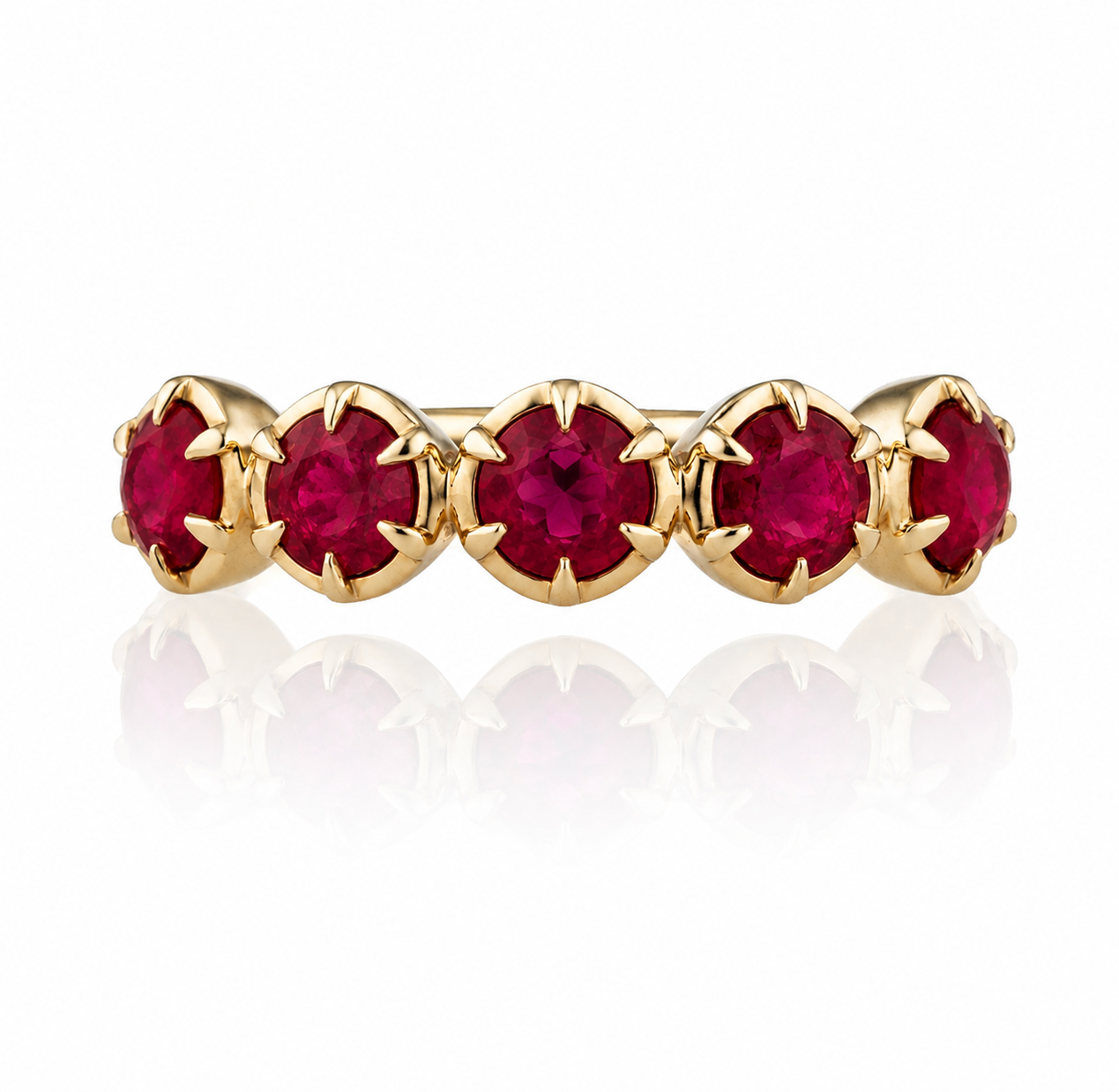 A gold eternity-style ring featuring five round-cut deep red gemstones in a prong setting, shown on a clean white background with a soft reflection below.