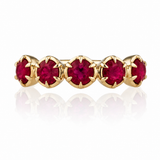 A gold eternity-style ring featuring five round-cut deep red gemstones in a prong setting, shown on a clean white background with a soft reflection below.