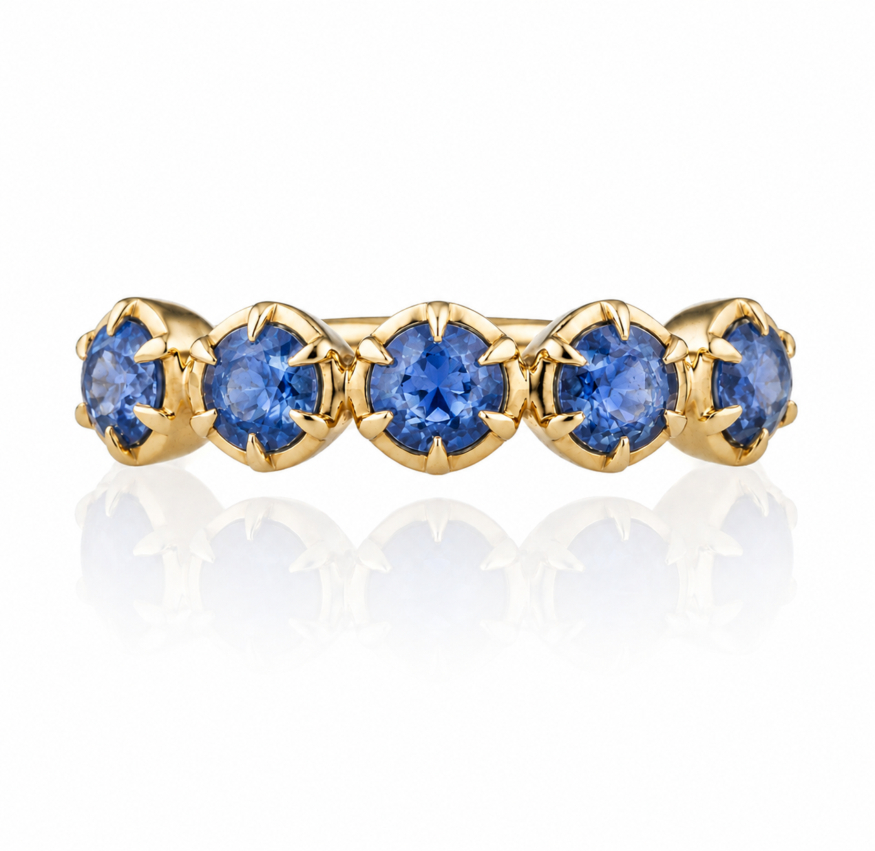 A yellow gold eternity ring featuring five round brilliant-cut blue gemstones in a row, set against a clean white background with a soft reflection below.