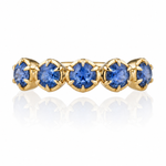 A yellow gold eternity ring featuring five round brilliant-cut blue gemstones in a row, set against a clean white background with a soft reflection below.
