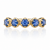 A yellow gold eternity ring featuring five round brilliant-cut blue gemstones in a row, set against a clean white background with a soft reflection below.