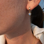 A close-up of a person's ear wearing a small gold huggie hoop earring with a round light blue gemstone, along with a delicate gold chain necklace.