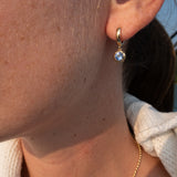 A close-up of a person's ear wearing a small gold huggie hoop earring with a round light blue gemstone, along with a delicate gold chain necklace.