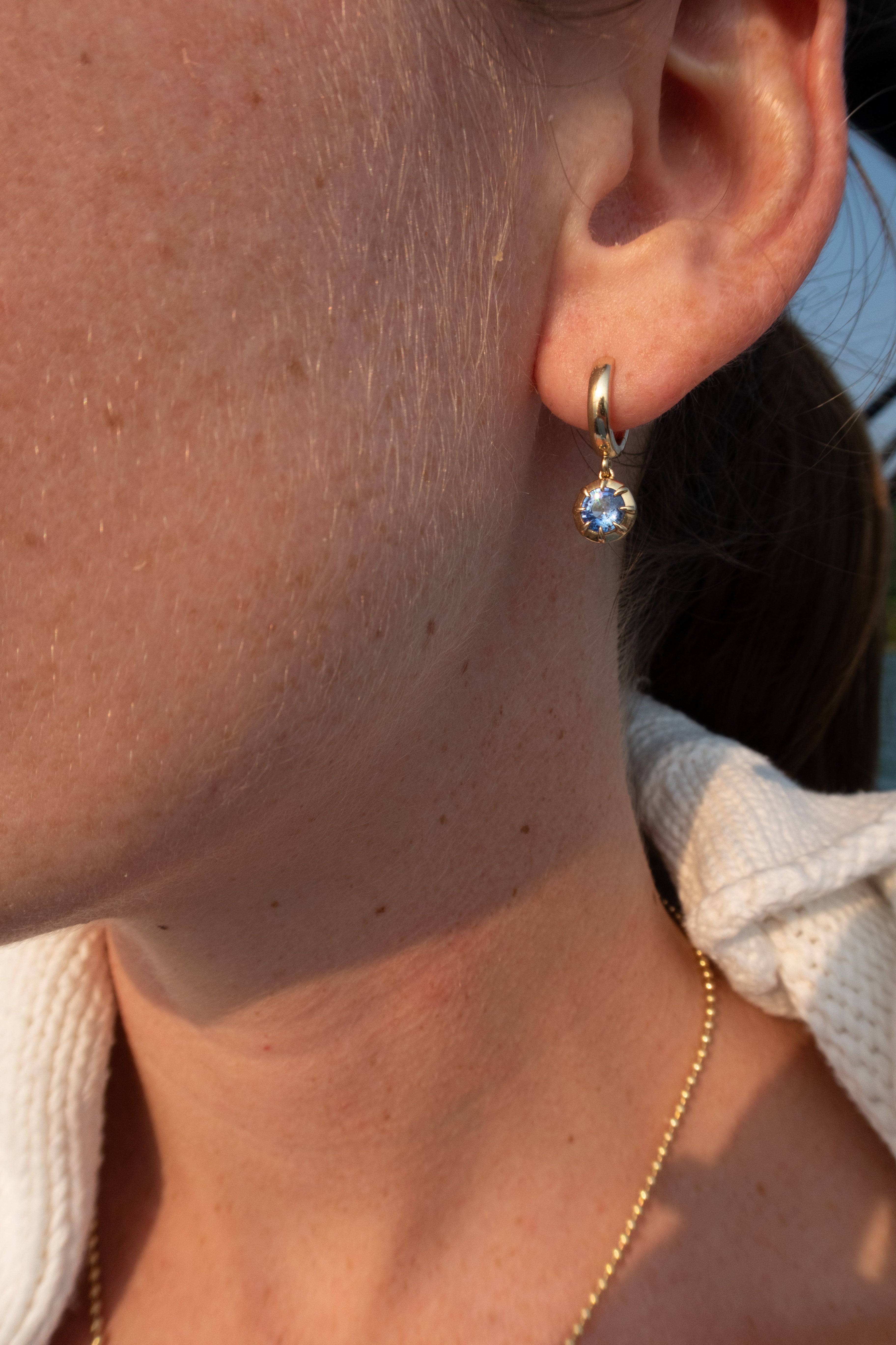 A close-up of a person's ear wearing a small gold huggie hoop earring with a round light blue gemstone, along with a delicate gold chain necklace.