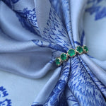 A close-up of a gold ring with five green emerald-cut stones, tied around a light blue and white patterned silk scarf.
