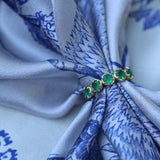 A close-up of a gold ring with five green emerald-cut stones, tied around a light blue and white patterned silk scarf.