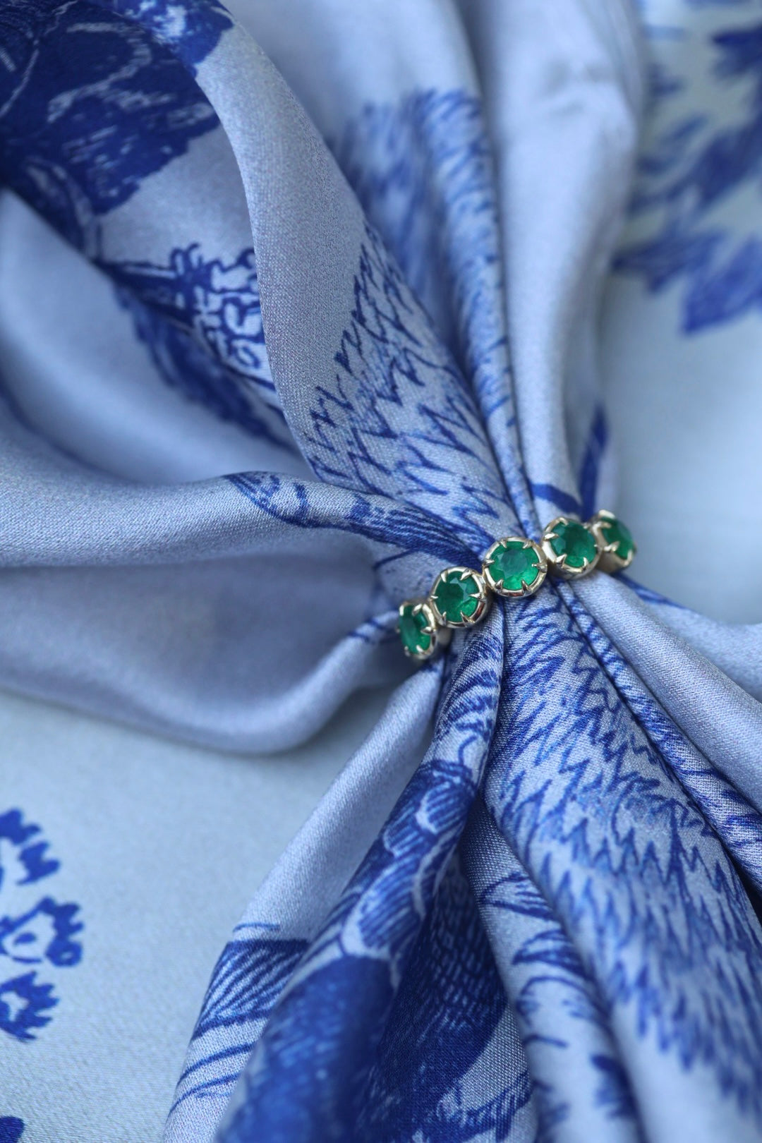 A close-up of a gold ring with five green emerald-cut stones, tied around a light blue and white patterned silk scarf.