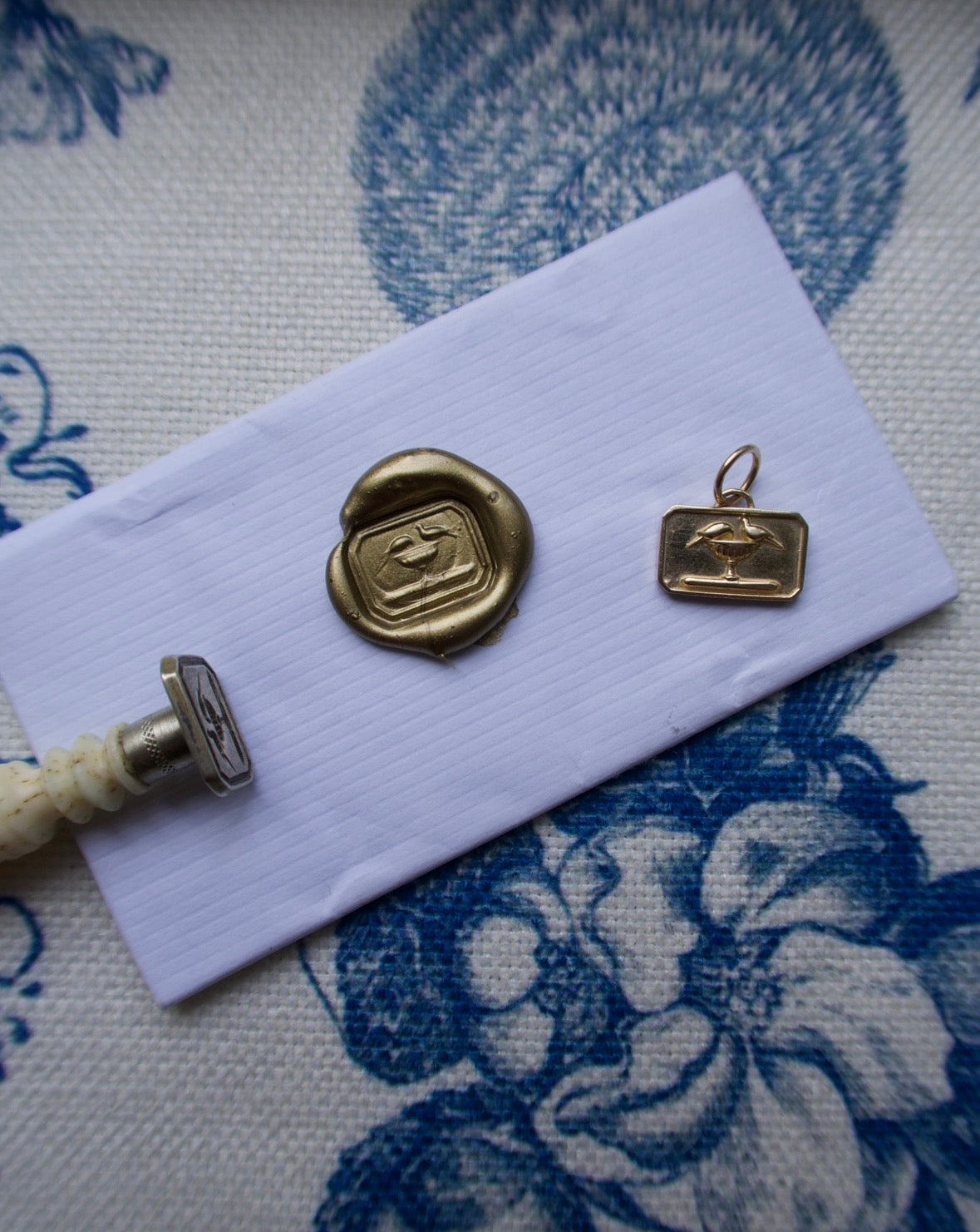 A small gold-toned metal charm or pendant with an engraved emblem, displayed on a white envelope alongside a wax seal and a small tool, all placed on a blue and white floral patterned fabric background.