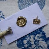 A small gold-toned metal charm or pendant with an engraved emblem, displayed on a white envelope alongside a wax seal and a small tool, all placed on a blue and white floral patterned fabric background.