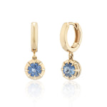 A pair of gold huggie hoop earrings with round sapphire gemstone drops, displayed on a clean white background with a soft reflection below.