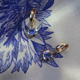 A pair of gold hoop earrings with round blue gemstones, displayed on a blue and white patterned silk fabric.