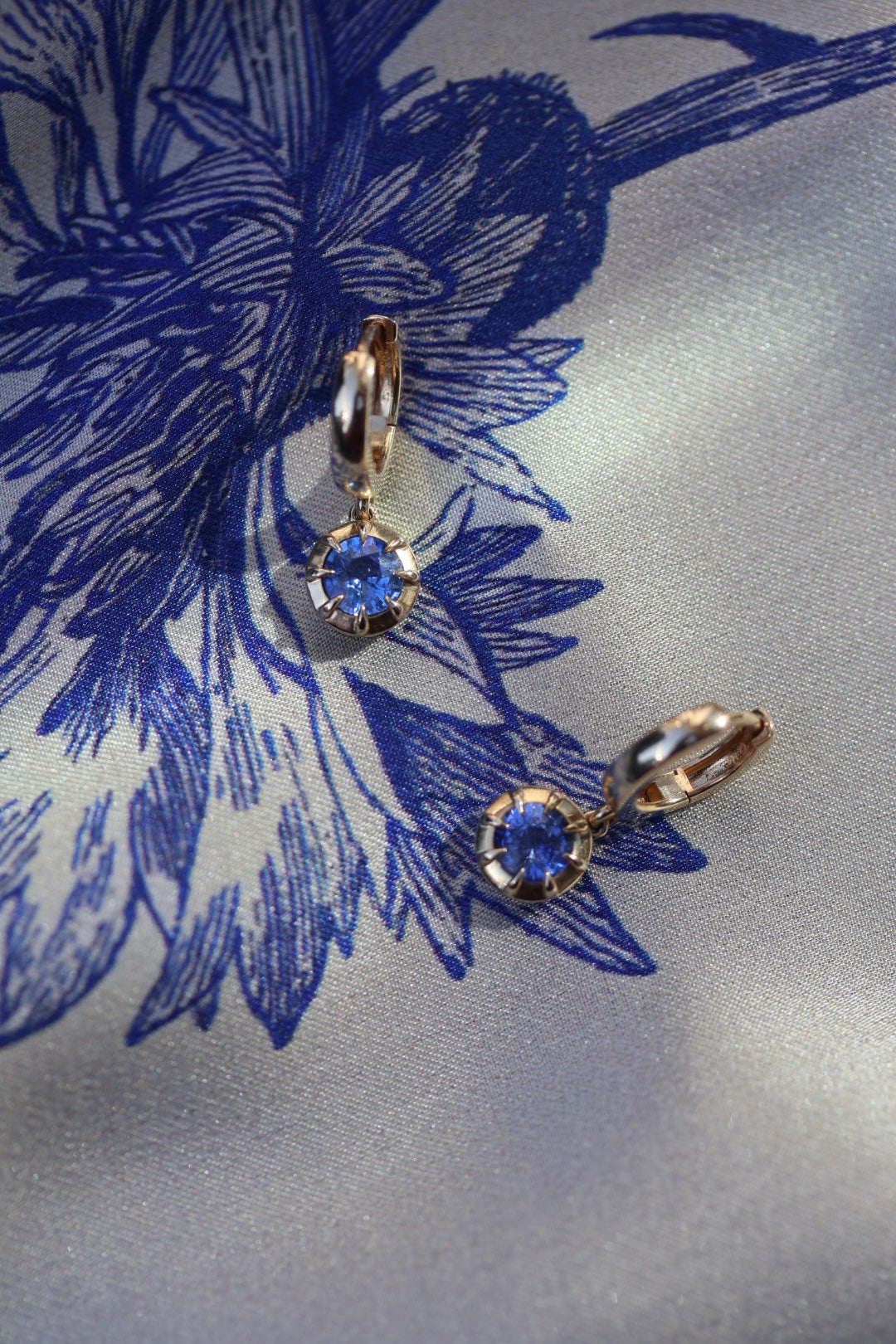 A pair of gold hoop earrings with round blue gemstones, displayed on a blue and white patterned silk fabric.
